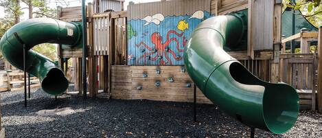 A playground with two green slides and a wooden climbing wall with a mural.