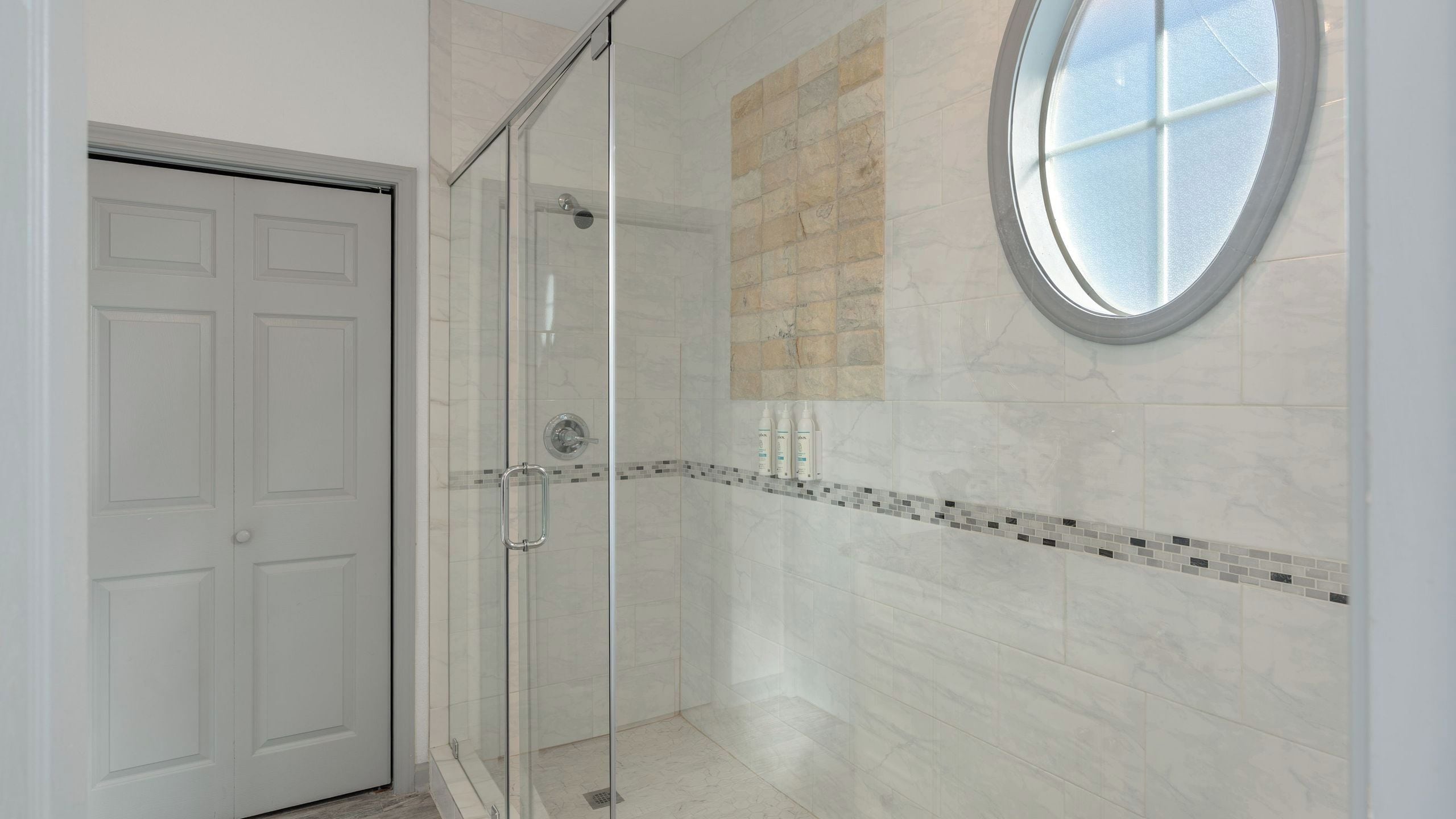 Une douche fermée par des parois de verre, avec un mur recouvert de carreaux, une fenêtre ronde et une porte.