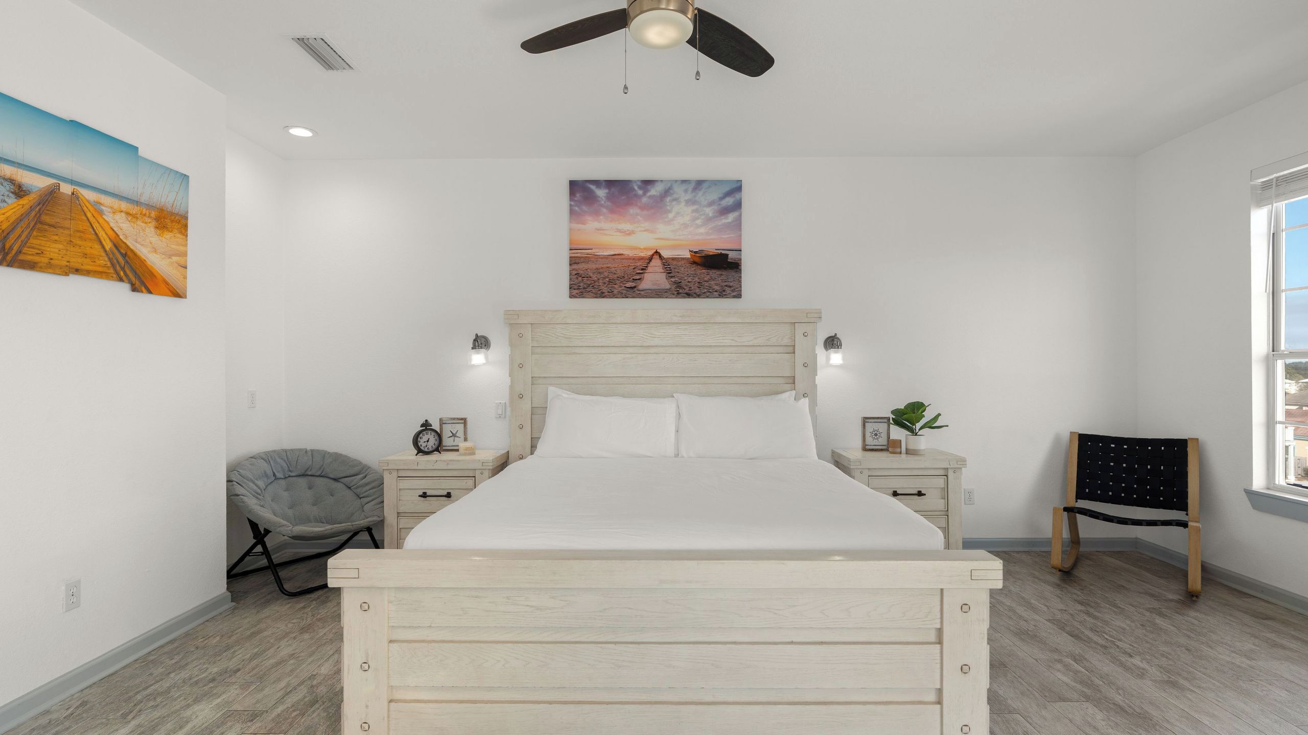 A bedroom with a wooden bed, a ceiling fan, a nightstand with a clock, a chair, and a window with a view.