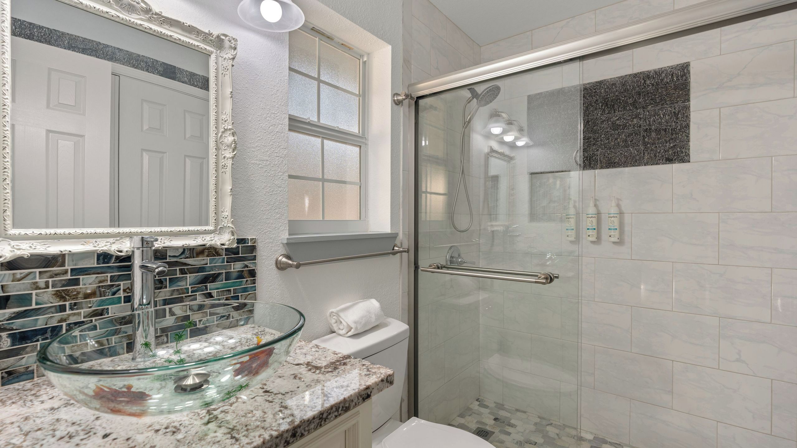 A bathroom with a glass sink, a shower with a glass door, a white toilet, and a mirror with an ornate frame.