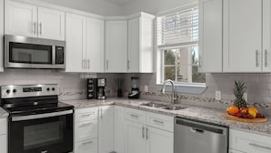 A modern kitchen with white cabinets, a built-in microwave, a stove, a coffee maker, and a blender. - Emerald Breeze by Avantstay Shared Pool + Tennis Court + Balcony Dining + Near Beach and Harbor Boardwalk (Destin)