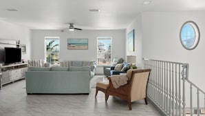 A spacious living room with a large flat-screen TV, a sofa, a chair, and a view of the outside through large windows. - Emerald Breeze by Avantstay Shared Pool + Tennis Court + Balcony Dining + Near Beach and Harbor Boardwalk (Destin)