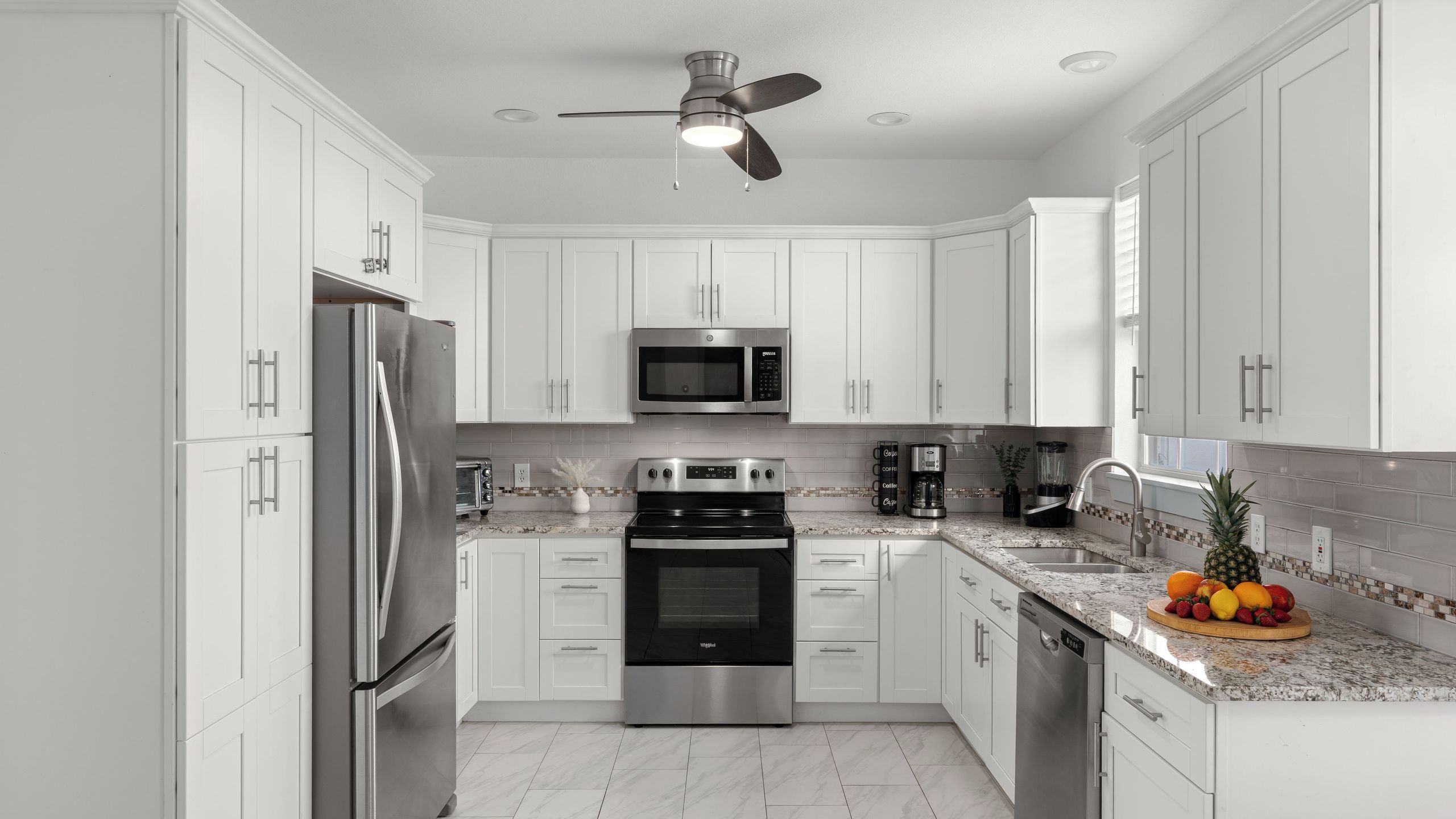 A modern kitchen with white cabinetry, stainless steel appliances, a ceiling fan, and a fruit bowl on the countertop.