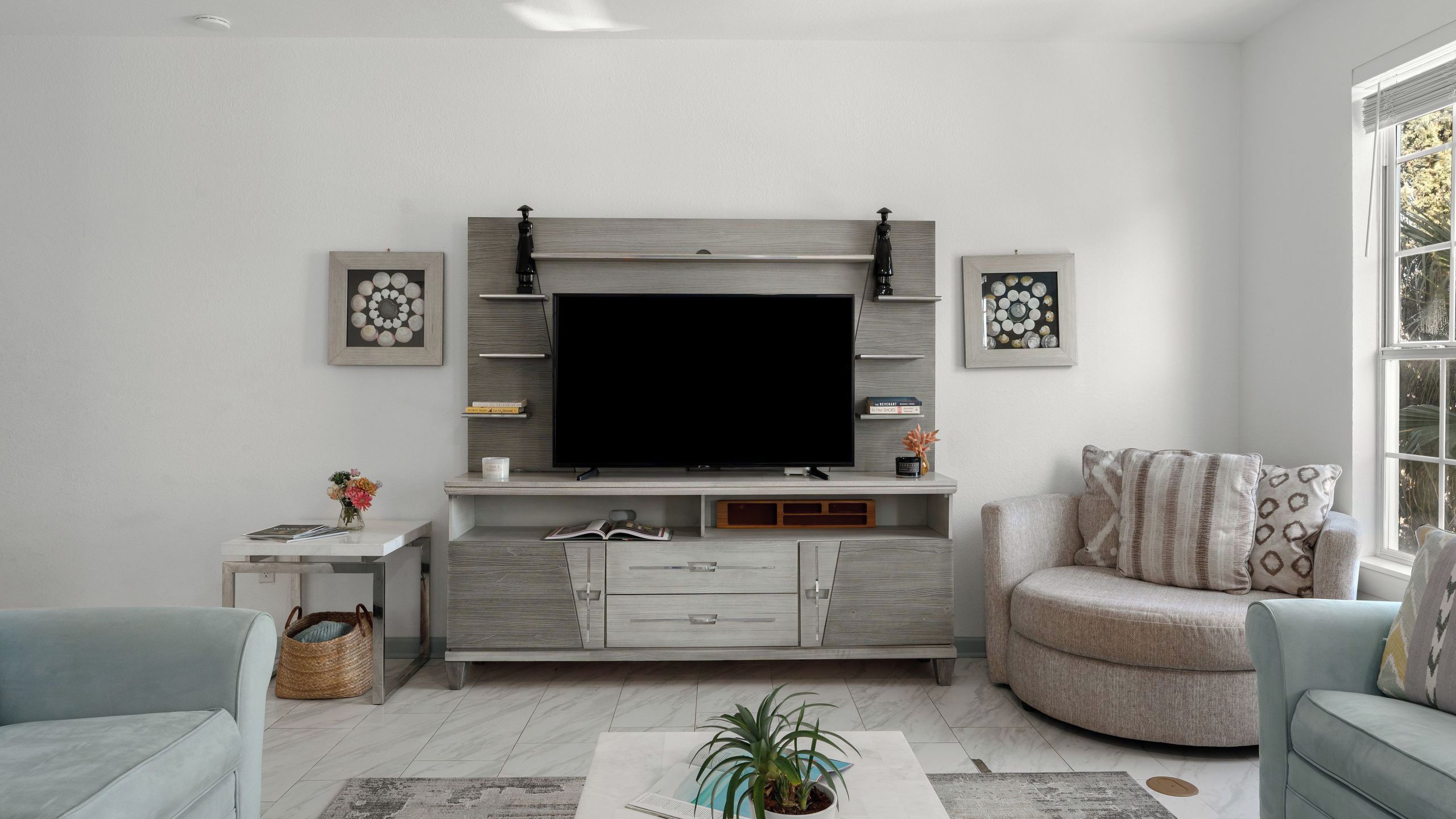 A living room with a flat-screen TV mounted on a grey wall, a sofa, a coffee table with a plant, and a window with a view.