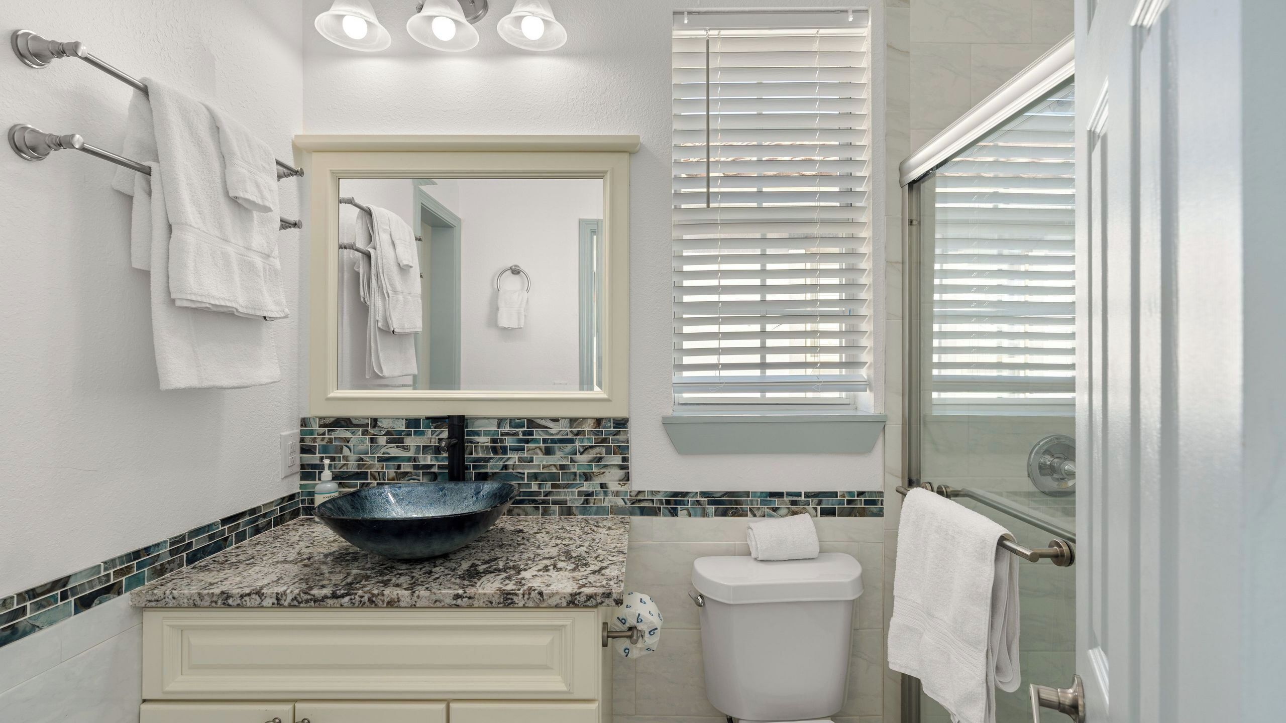 A bathroom with a marble countertop, a vessel sink, a toilet, a glass shower enclosure, and a mirror with a towel rack.