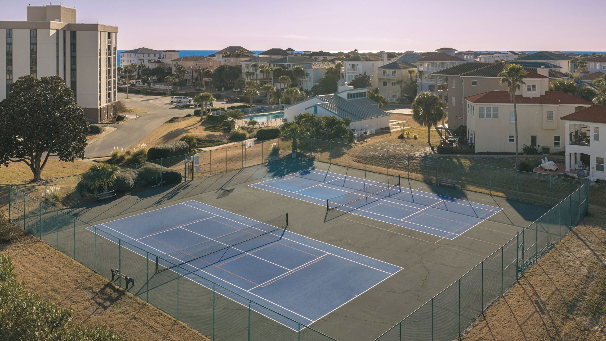 Una cancha de tenis rodeada de edificios residenciales, un área de estacionamiento y un árbol grande.