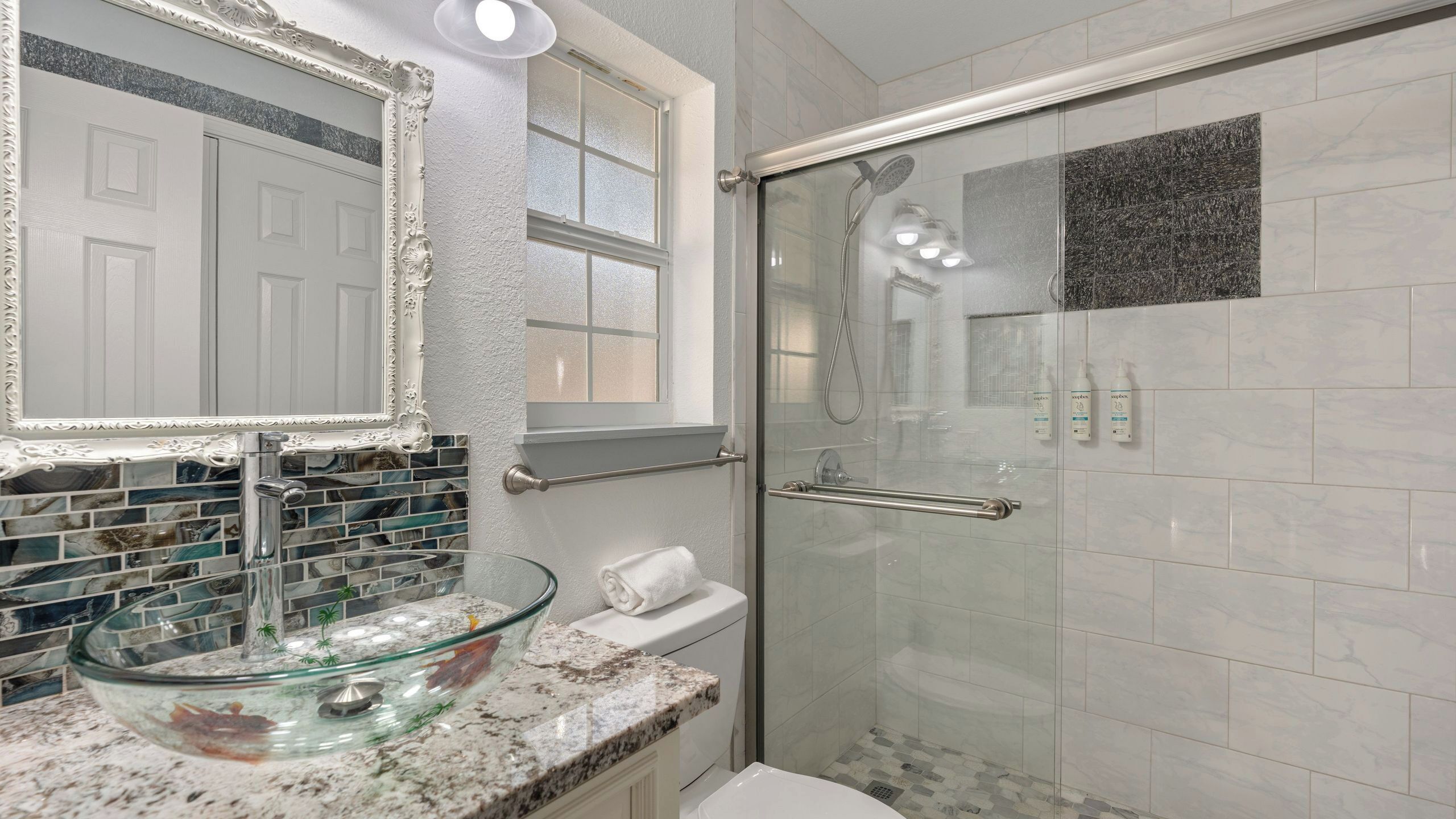 A bathroom with a glass sink, a shower with a glass door, a white toilet, and a mirror with an ornate frame.