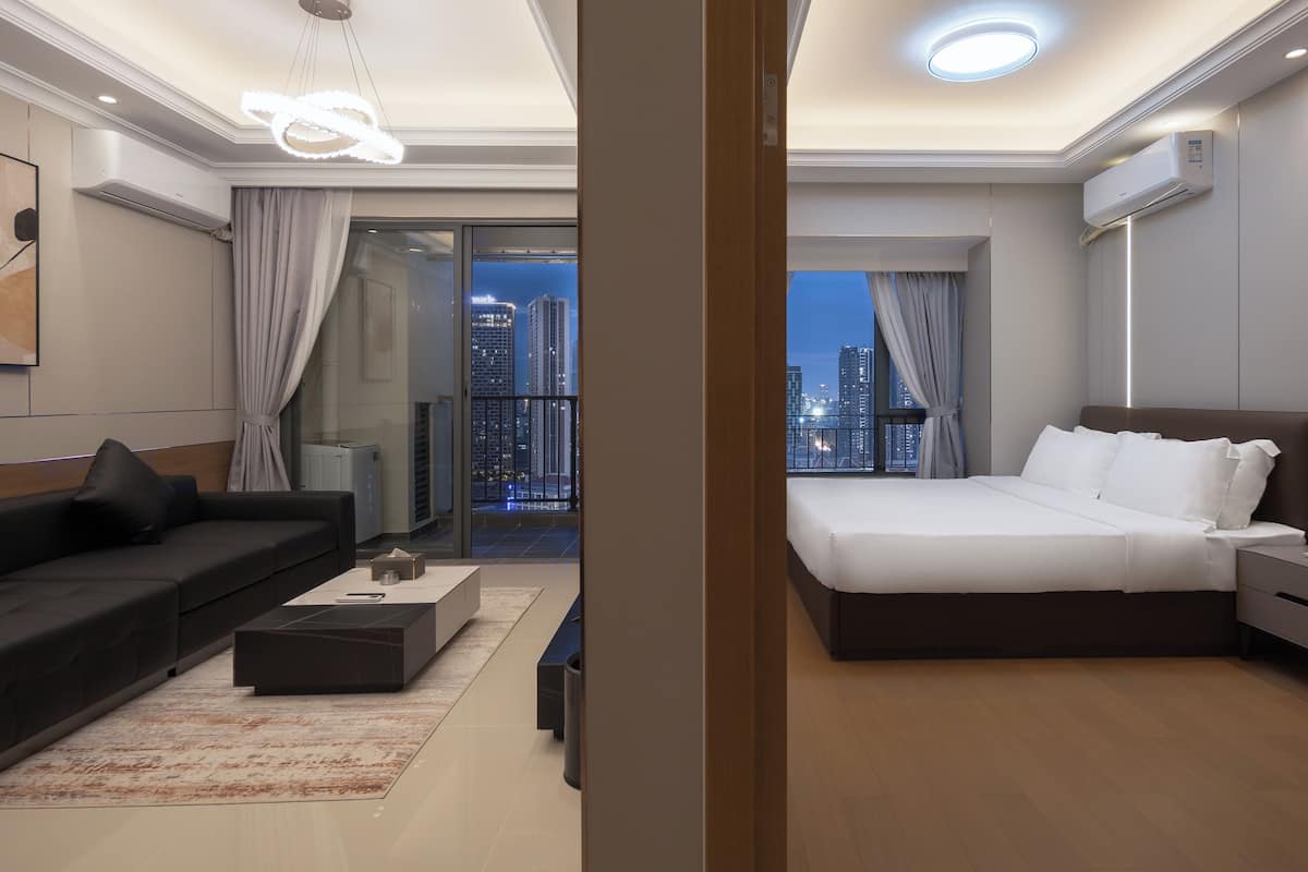 A modern hotel room with a large bed, a sofa, a coffee table, and a view of the cityscape at night.