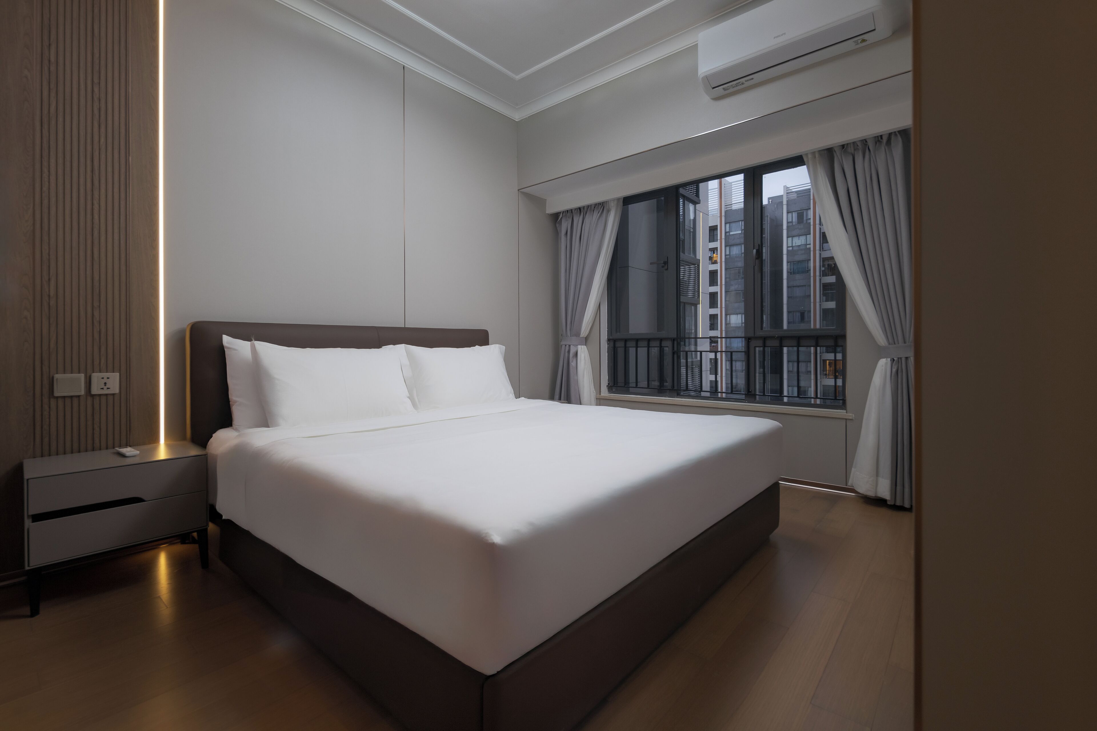 A modern hotel room with a large bed, a bedside table, a nightstand, a window with curtains, and an air conditioning unit.