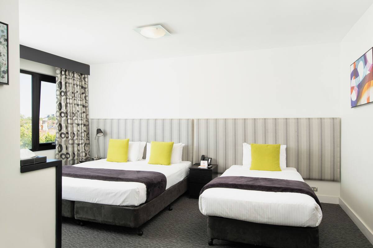 Habitación de hotel con dos camas, un escritorio, una silla, una ventana con cortinas, una mesita de noche y un cuadro en la pared.