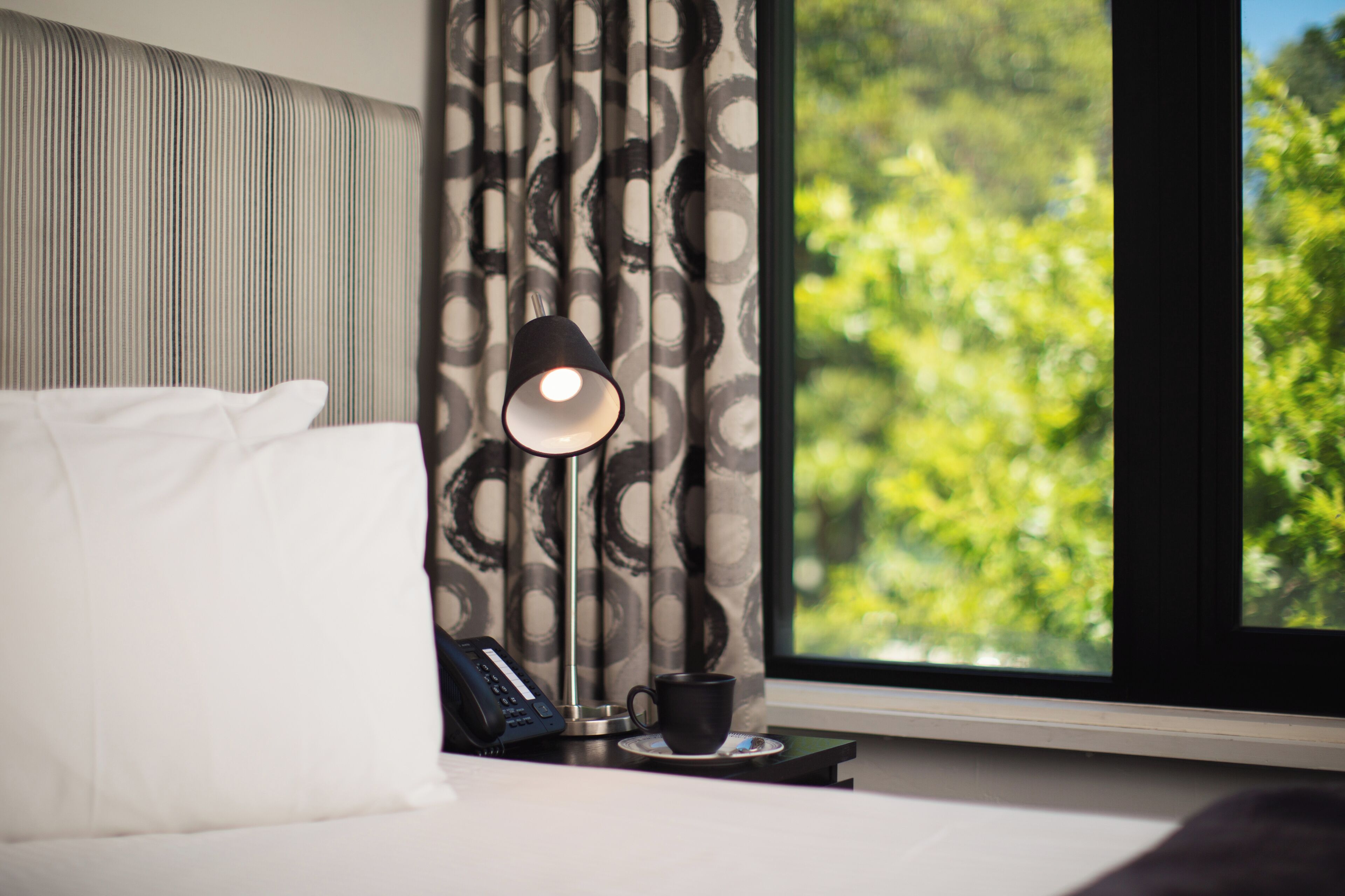 A neatly made bed with white linens, a black telephone, a black cup, and a floor lamp on a bedside table. A window with a view of greenery is visible.