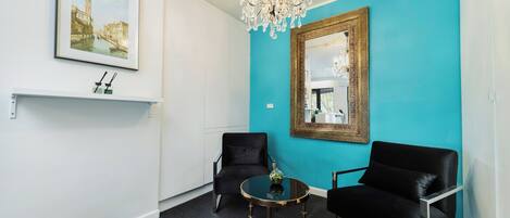 A room with teal walls, a chandelier, two black chairs, a round table, and a framed painting.