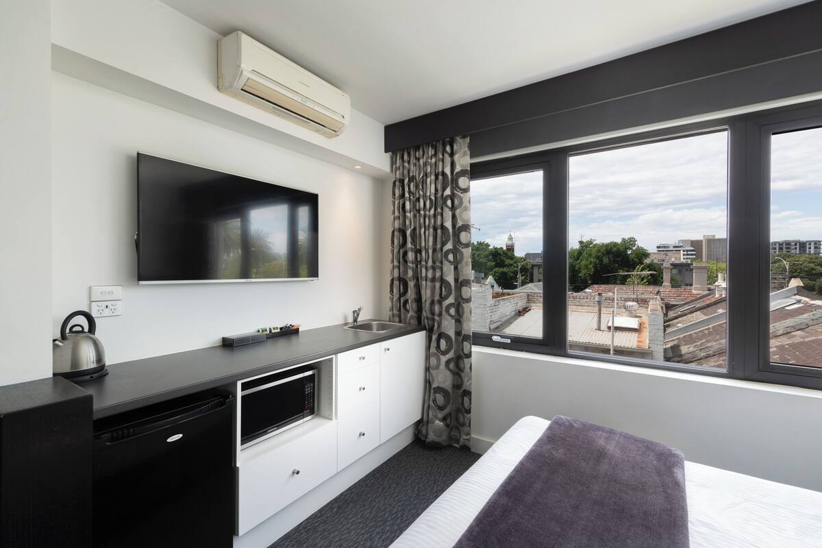 Una habitación de hotel moderna con una televisión de pantalla plana, una zona de cocina con microondas, un lavabo y un amplio ventanal con vistas a la ciudad.