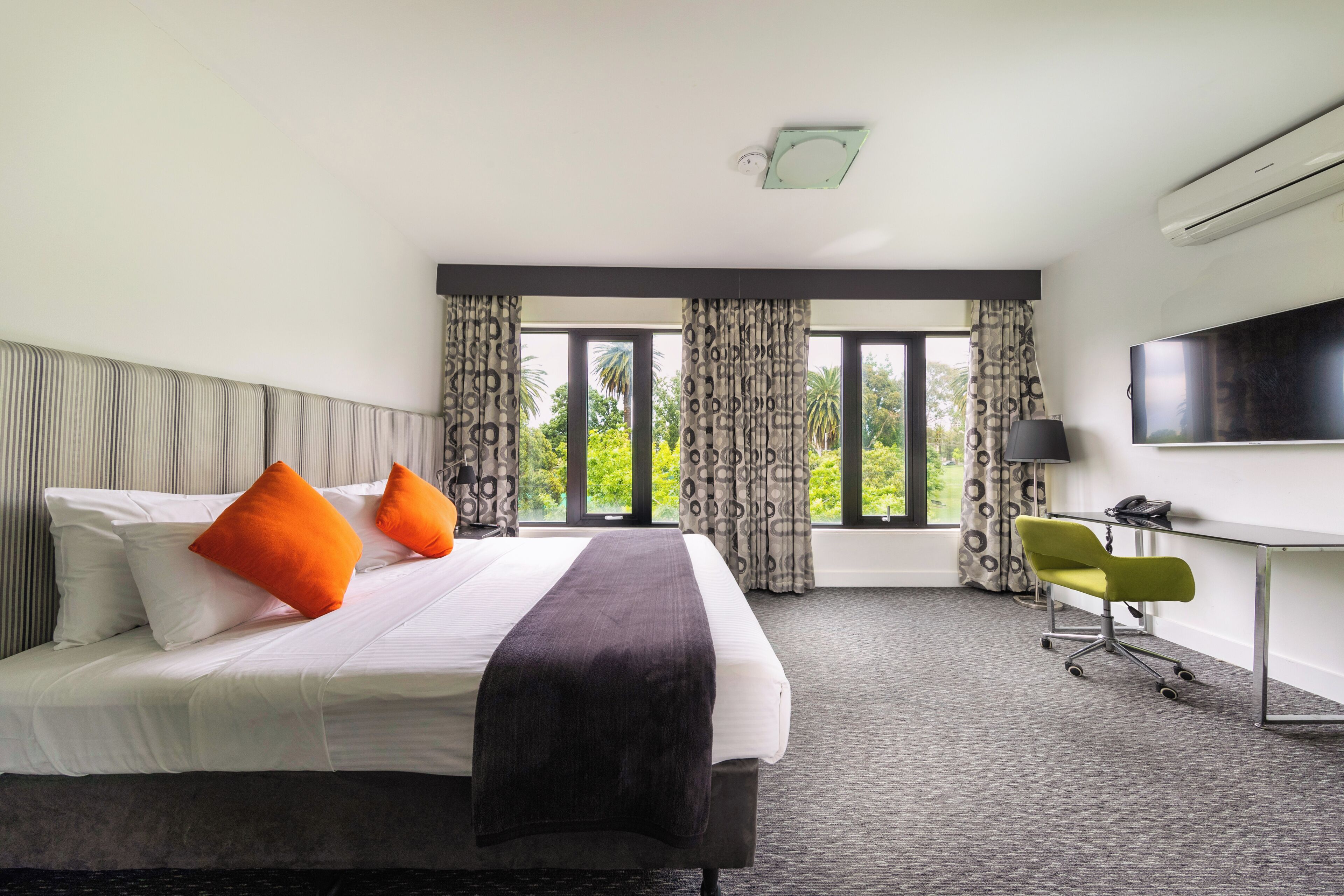 Habitación de hotel con cama, escritorio, silla, televisor y ventanas amplias con cortinas.