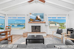 A spacious living room with a fireplace, a large painting of a coastal scene, and a view of the sea and boats through large windows. - Bayfront 5BR with Stunning Harbor Views, Only Steps to the Beach! (Newport Beach)