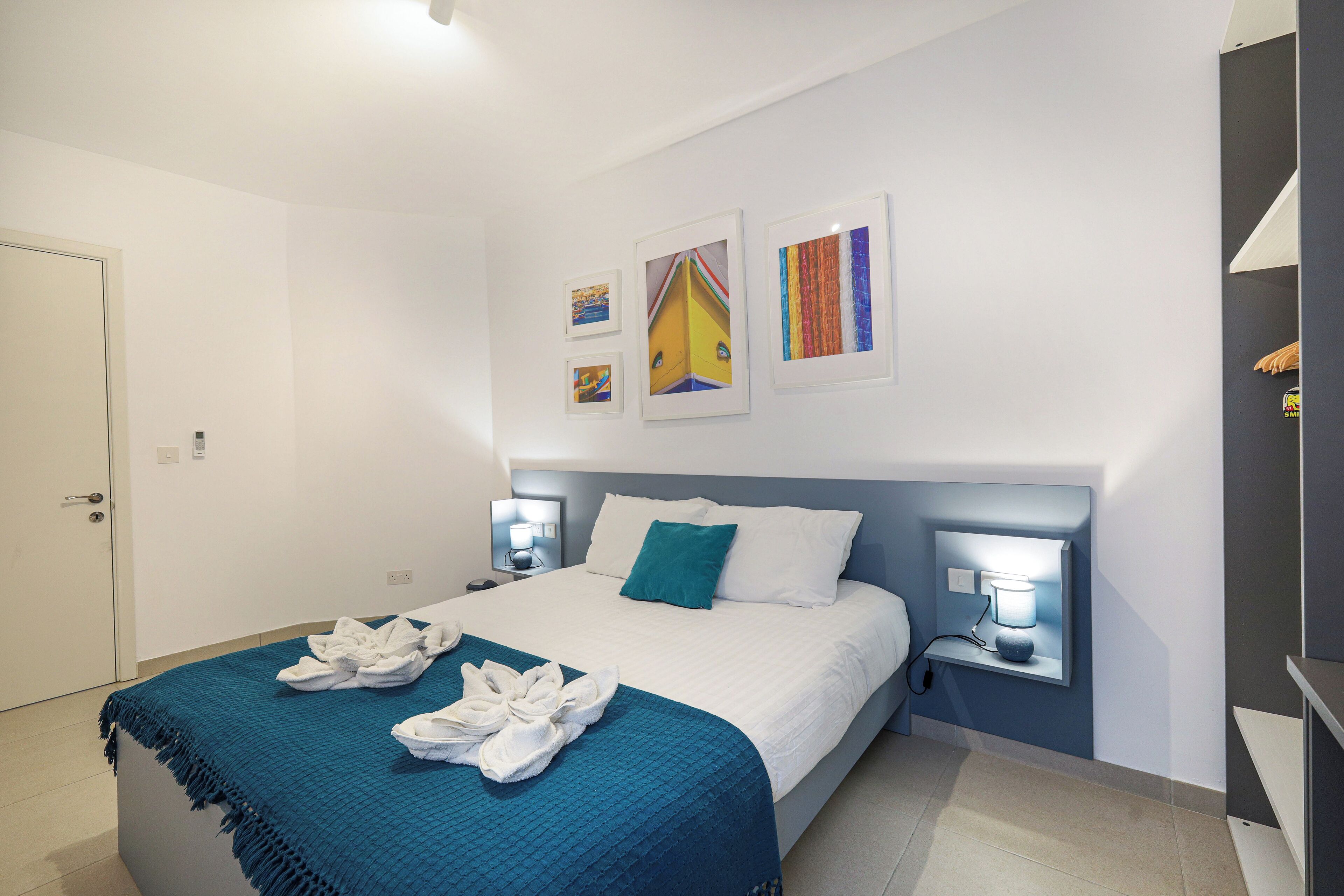 Una camera d'albergo con un letto, due comodini con lampade, una coperta blu e un quadro appeso al muro.