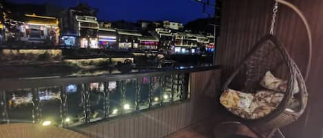 Un balcón con una silla colgante y vistas a una zona iluminada a la orilla del río al anochecer.