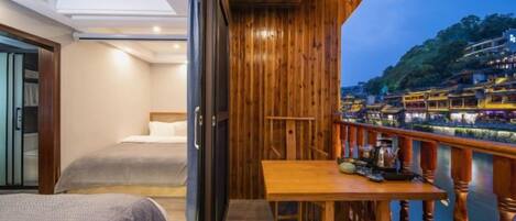 A balcony with a wooden railing overlooking a river, a bed with a gray bedspread, and a table with various items.