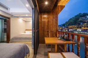 Een balkon met houten hekwerk met uitzicht op een rivier, een bed met een grijze sprei, en een tafel met diverse voorwerpen.