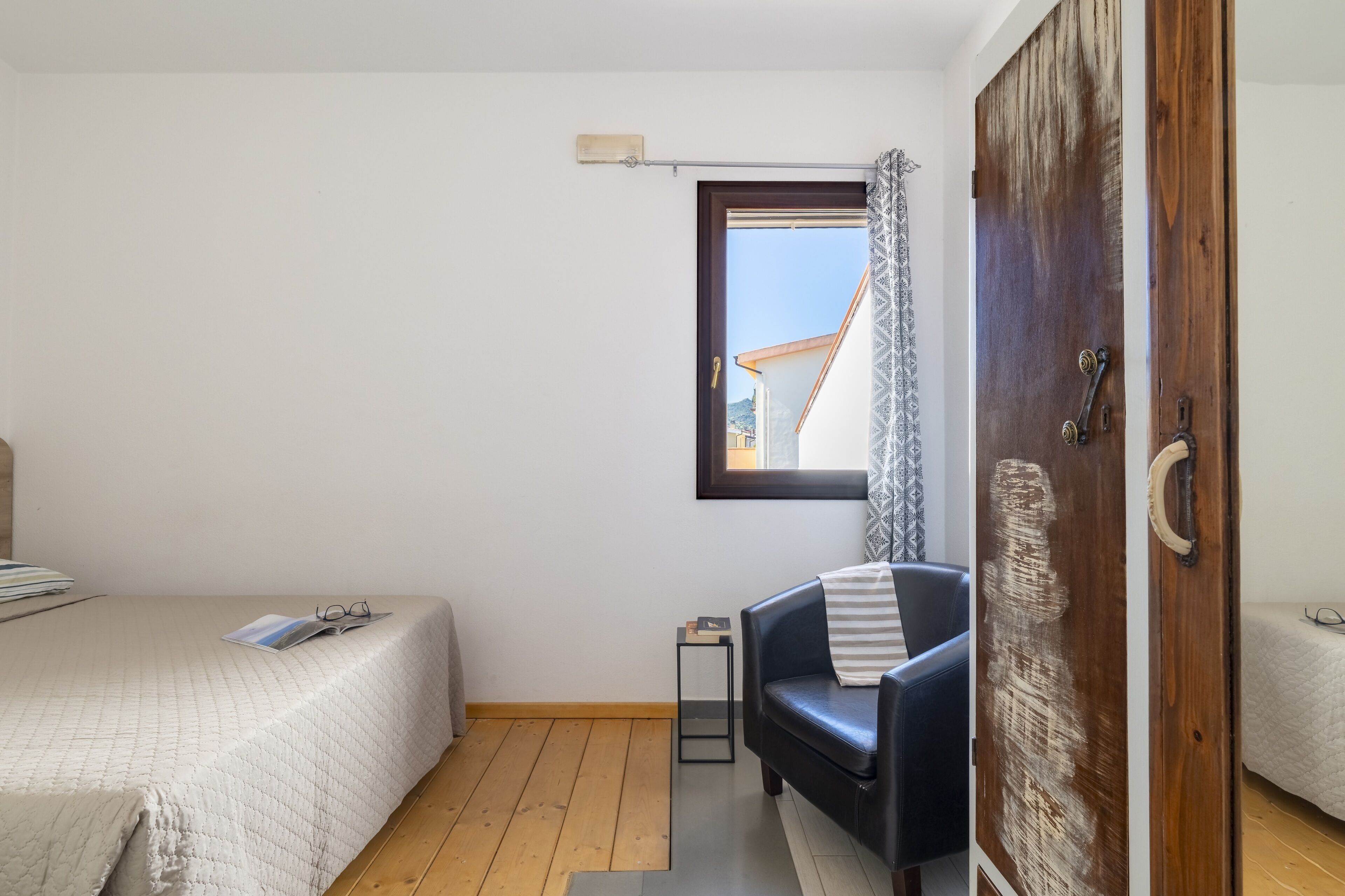 Quarto com cama, uma cadeira, uma mesinha e janela com vista para edifícios.