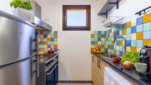 Cocina compacta con refrigerador, estufa, horno y lavadora. Una ventana con marco de madera se encuentra sobre el lavamanos, y azulejos coloridos decoran el salpicadero.
