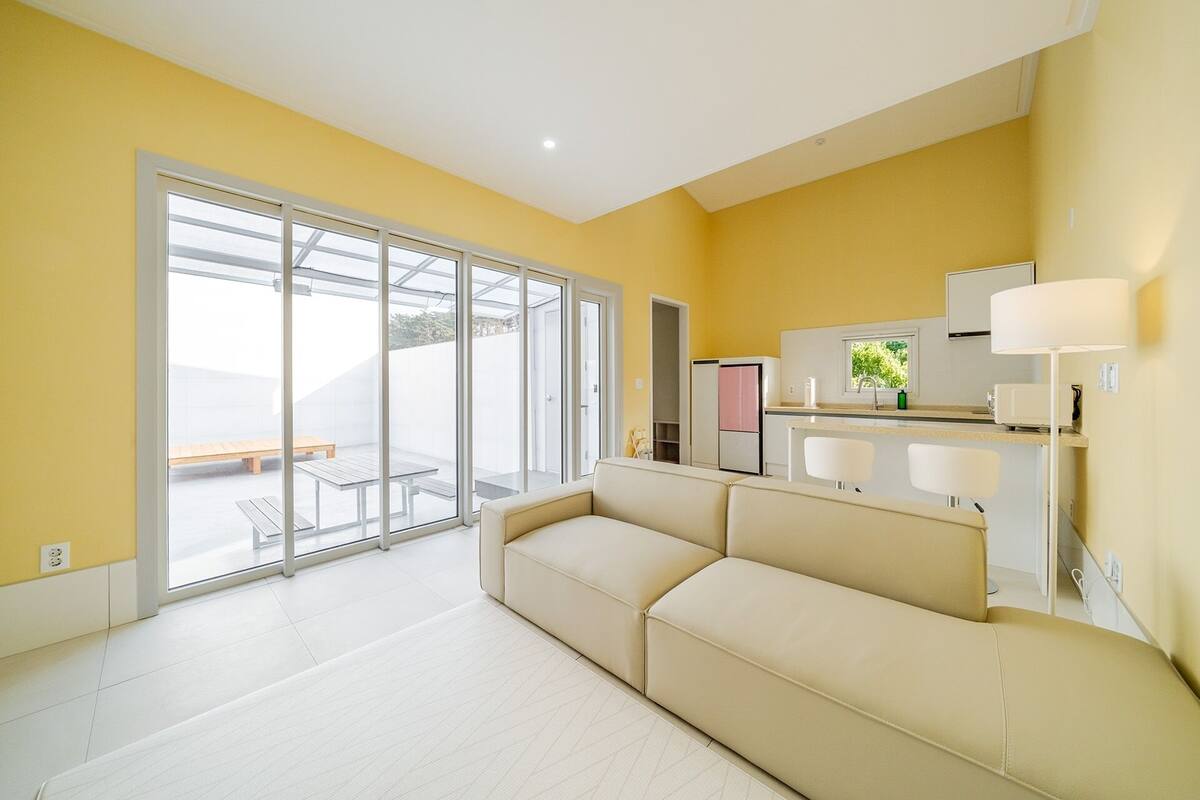 Una sala de estar moderna con un sofá, una cocina con refrigerador y fregadero, y un ventanal corredizo que da a un patio exterior.
