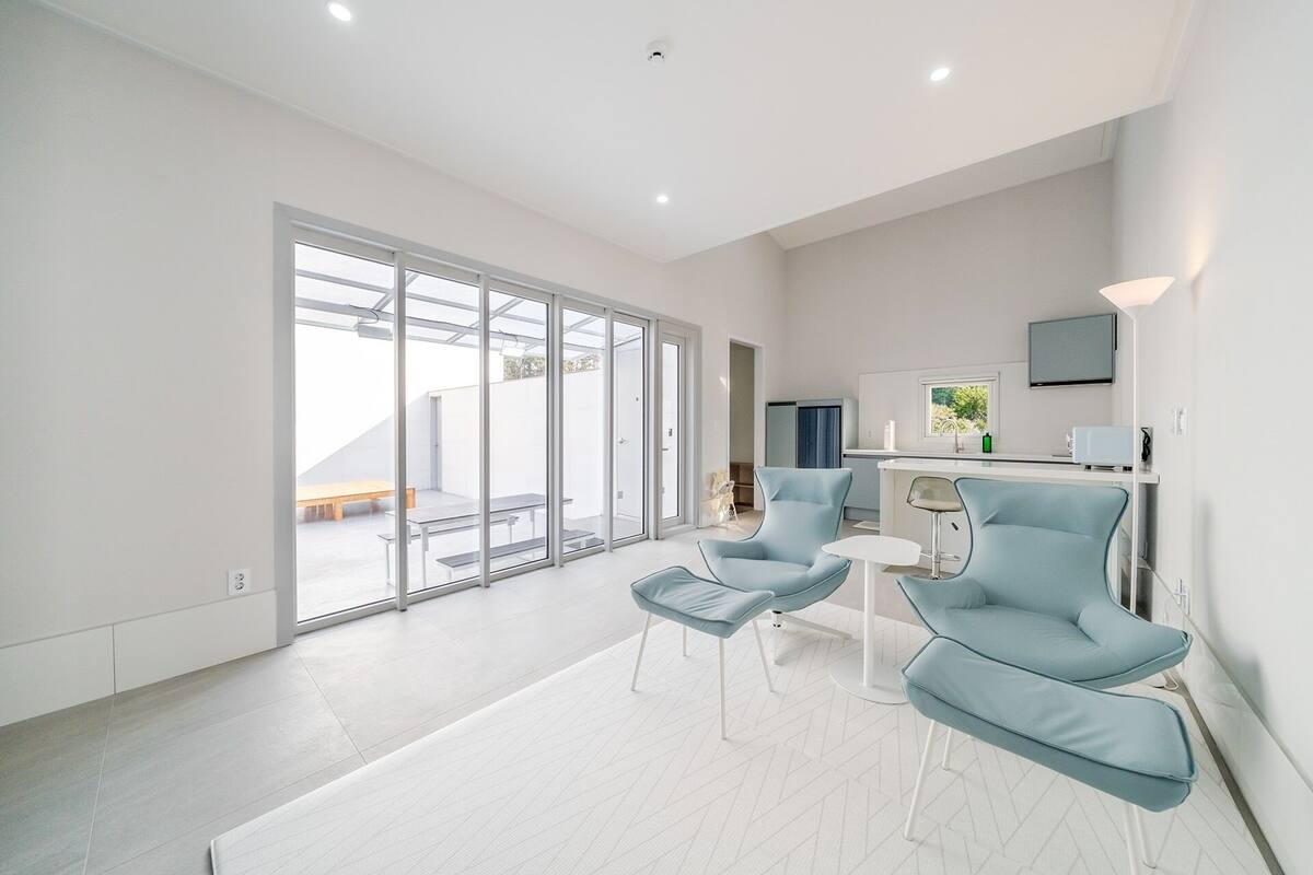 Un espacio habitable moderno y amplio, con un ventanal corredero que da a un patio exterior, una zona de cocina con televisor de pantalla plana, y un rincón de descanso con dos sillas azules y una mesita.