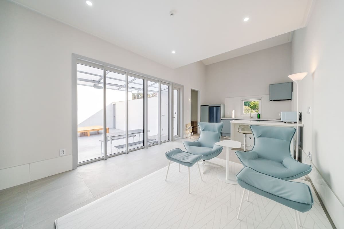 Un espace de vie moderne et spacieux, doté d’une porte-fenêtre coulissante donnant sur une terrasse extérieure, d’un coin cuisine avec un téléviseur à écran plat et d’un coin salon avec deux fauteuils bleus et une petite table.