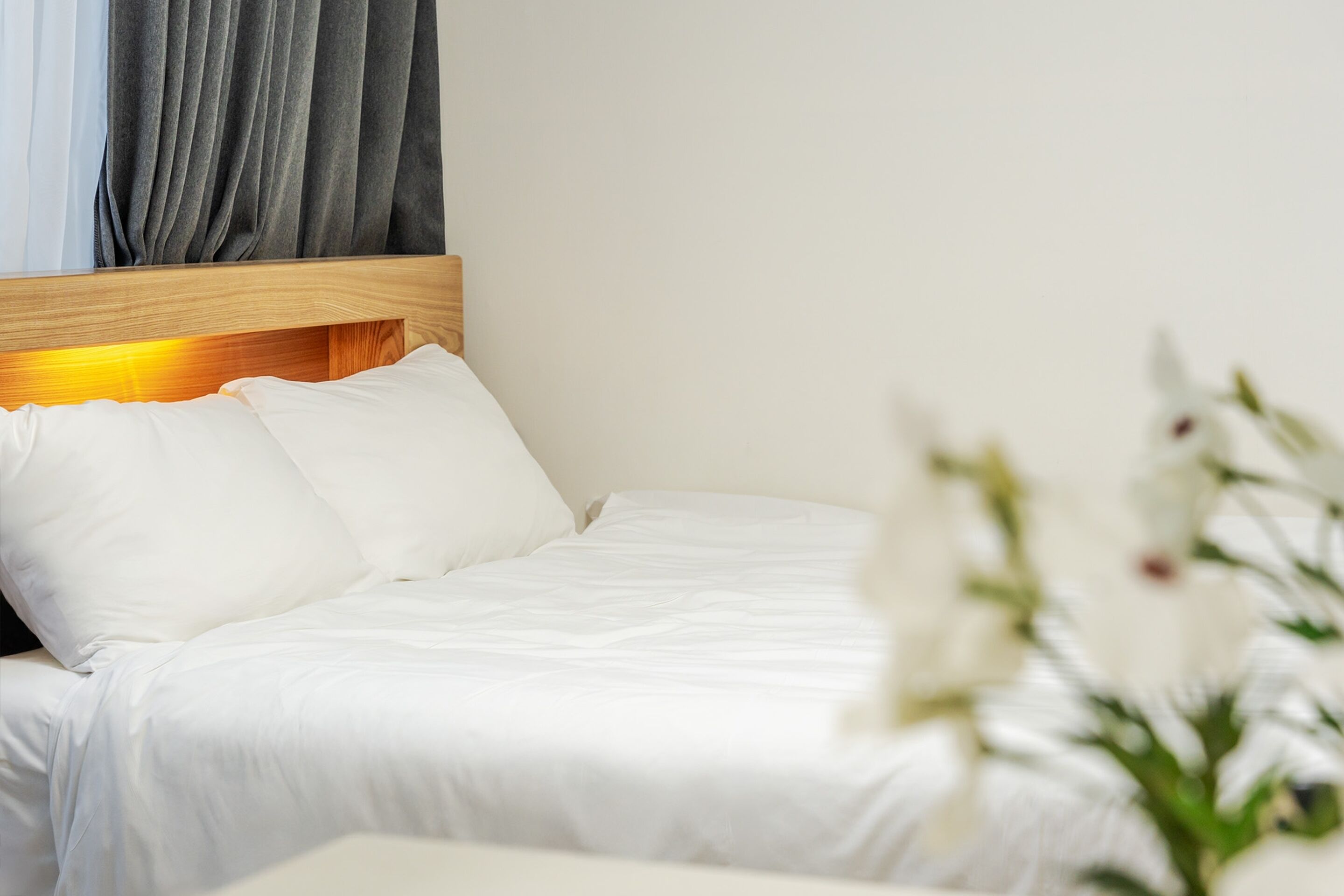 Una cama bien tendida con sábanas y almohadas blancas, un cabecero de madera y una ventana con cortinas grises.