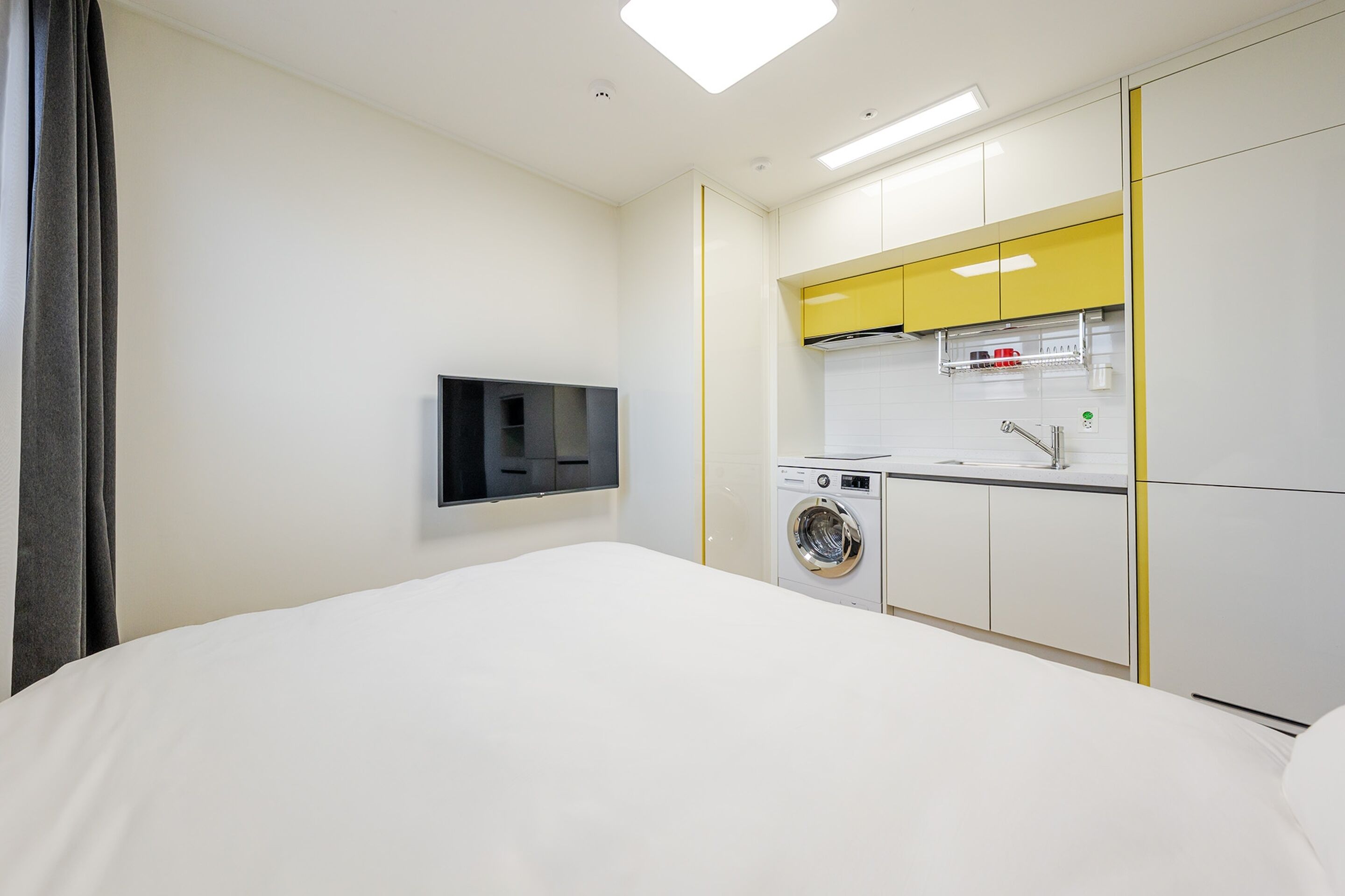 A hotel room with a bed, a washing machine, a sink, and a TV mounted on the wall.