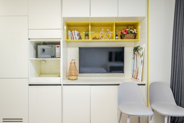 A modern room with a TV, white chairs, a yellow shelf with books and decorative items, and a window with a view of palm trees. - Yangyang Oceanstay 24K (Yangyang)