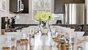 A dining table set with glasses and plates, a vase with flowers, and modern kitchen appliances in the background. - 9BR Table Rock Lake Lodge w/ Hot Tub & Game Room • Near SDC (Branson)