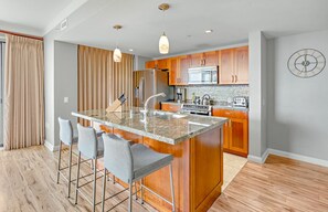 A modern kitchen with a central island, bar stools, wooden cabinets, stainless steel appliances, and pendant lighting. - Maui Resort Rentals: Honua Kai Hokulani 413/5 – Luxe 3BR Oceanview Escape! (Lahaina)