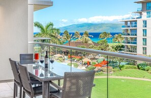 A balcony with wicker furniture, a glass table, and a view of a resort area with palm trees and a pool. - Maui Resort Rentals: Honua Kai Hokulani 413/5 – Luxe 3BR Oceanview Escape! (Lahaina)