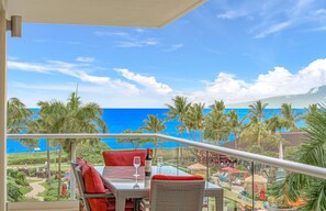 A balcony with a table and chairs, overlooking a beach with palm trees and a clear blue sky. - Maui Resort Rentals: Honua Kai Hokulani 413/5 – Luxe 3BR Oceanview Escape! (Lahaina)