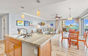 A modern kitchen with granite countertops, a built-in dishwasher, and a dining area with wooden chairs and a glass table. - Maui Resort Rentals: Honua Kai Hokulani 413/5 – Luxe 3BR Oceanview Escape! (Lahaina)