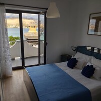 Egy hálószoba, amelyben található egy ágy, egy éjjeliszemélyzet, egy lámpa, egy ablak, ahonnan egy épületre és egy víztömegre nyílik kilátás.
