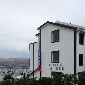 A white building with 'HOTEL BISER' signage, a visible staircase, and a view of a distant town. - Biser Hotel (Pag)