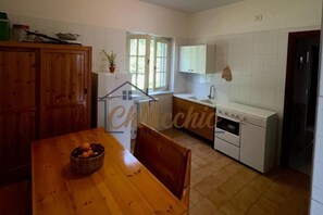 A kitchen with white cabinets, a wooden table with a fruit bowl, and a window providing natural light. - Villetta Gentili - ChiacchioProperty (Policastro Bussentino)
