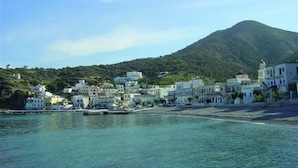 A coastal town with buildings along the waterfront, a mountain in the background, and clear blue water. - Glaros Studios (Karpathos)