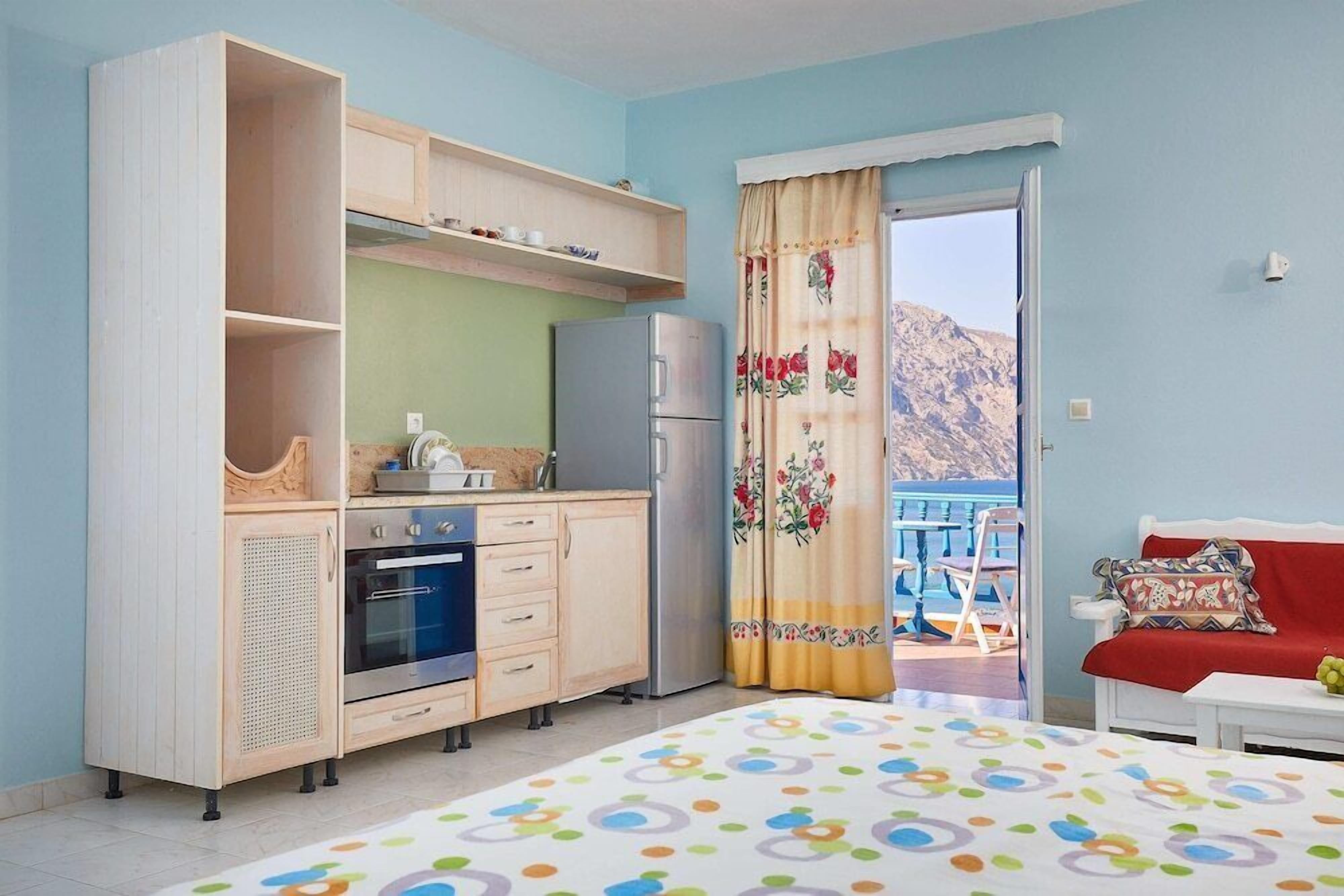 A bedroom with a bed, a kitchenette, a balcony with a view of the mountains, and a window with curtains.