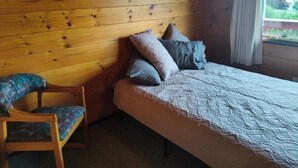 Une pièce aux murs lambrissés, comprenant un lit, des oreillers, une chaise et une fenêtre donnant sur un paysage agréable.