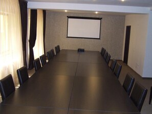 Een vergaderzaal met een lange tafel en stoelen, een projectiescherm en gordijnen.
