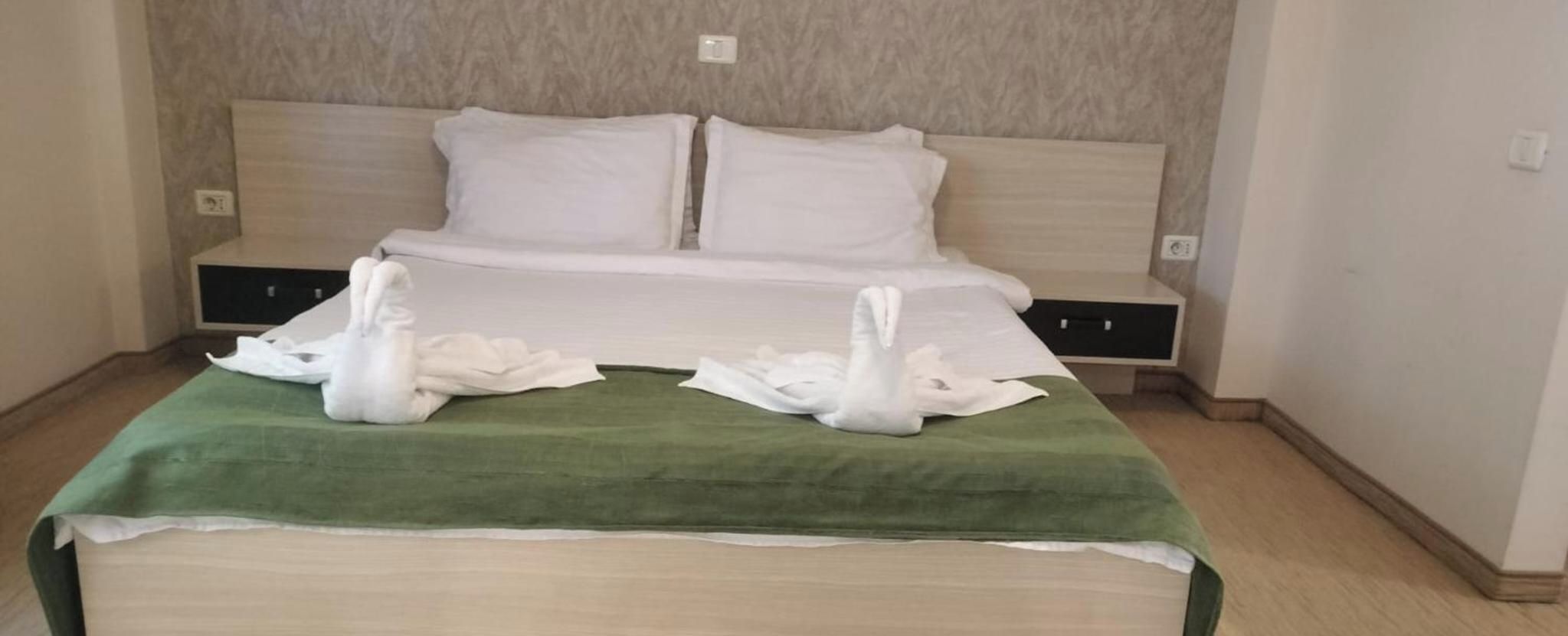 A hotel room with a bed, two pillows, a green bedspread, and two white towels arranged on the bed.