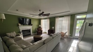 Ein Wohnzimmer mit Ecksofa, Kamin, Deckenventilator und großem Fenster mit Jalousien.