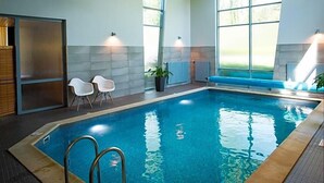 Una piscina interior con paredes de vidrio transparente, agua azul y una zona de estar con sillas blancas.