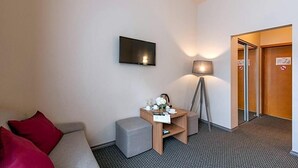 Una habitación de hotel con un sofá, una mesita con lámpara, un televisor montado en la pared y una puerta con un cartel de prohibido fumar.