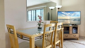 A dining area with a glass table, wooden chairs, a vase with flowers, a large mirror, a flat-screen TV, and a cabinet with a remote control. - 2BR Condo at Fantasy Island – Pool, Beach , Bikes (Englewood)
