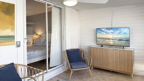 Una habitación con puerta corrediza de vidrio que da a un dormitorio, ventilador de techo, televisor sobre un mueble de madera y una silla de mimbre.