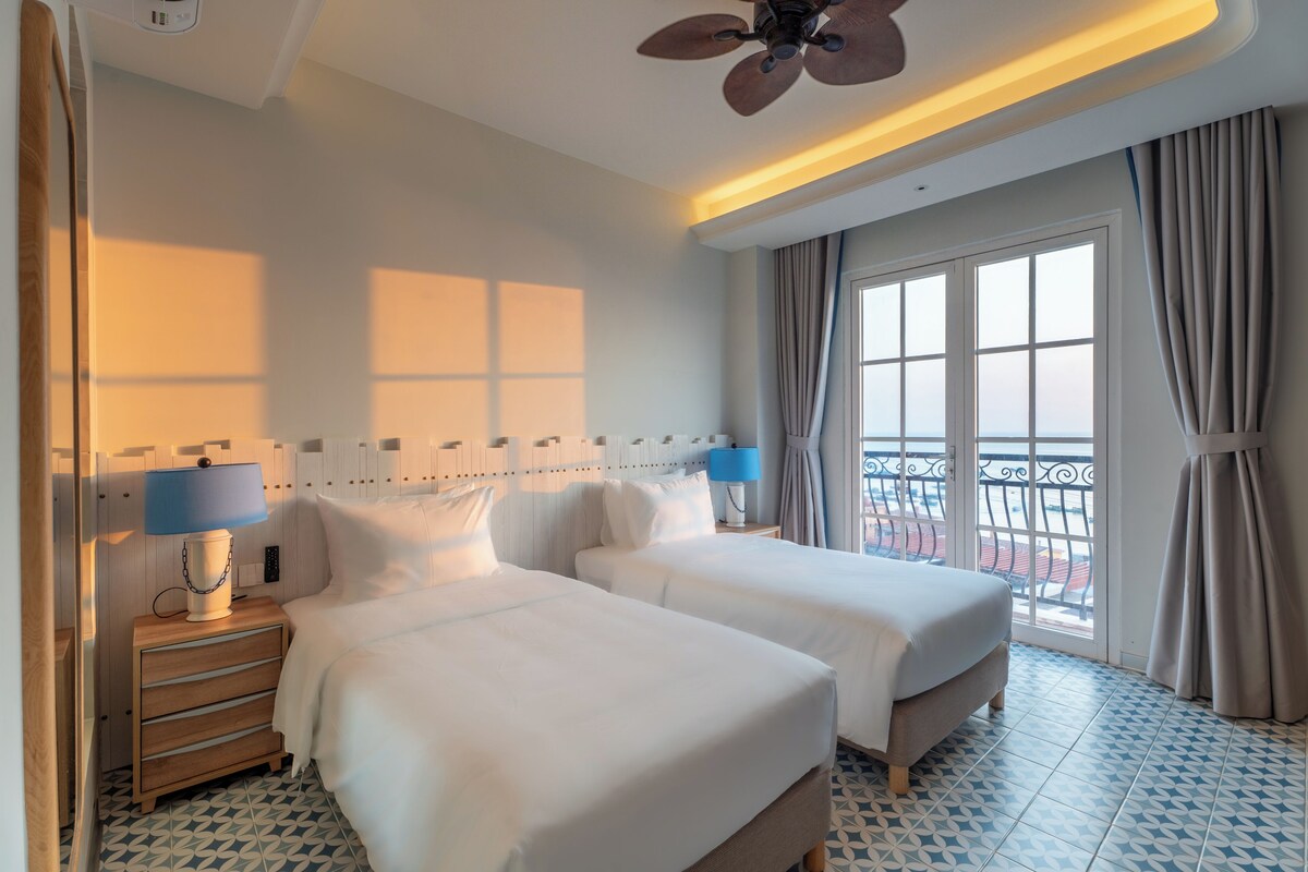 A hotel room with two beds, a ceiling fan, a window with curtains, and a balcony view.